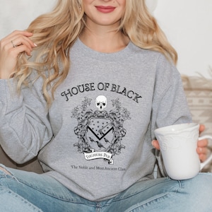 May include: Grey sweatshirt with a black graphic of a coat of arms. The graphic features a skull, ravens, and the text "House of Black", "Toujours Pur", and "The Noble and Most Ancient Clan".