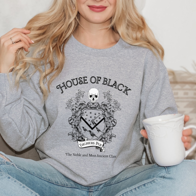 May include: Grey sweatshirt with a black graphic of a coat of arms. The graphic features a skull, ravens, and the text "House of Black", "Toujours Pur", and "The Noble and Most Ancient Clan".