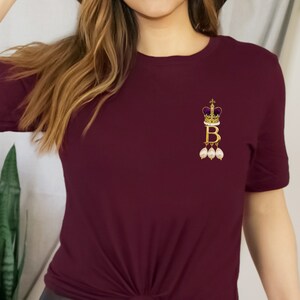 Może przedstawiać: Bordowy t-shirt z złotą koroną i literą "B" z wiszącymi perłami.