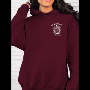 May include: A maroon hoodie with a front pocket and a hood. The upper left chest area features a white graphic with the words "HOUSE OF BLACK" and other text. The hoodie is made of a soft material.