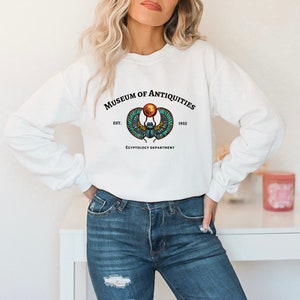 Puede incluir: Sudadera blanca con un estampado gr&aacute;fico de un escarabajo con alas y un disco solar. El texto "Museum of Antiquities Est. 1925 Egyptology Department" est&aacute; impreso debajo del gr&aacute;fico.