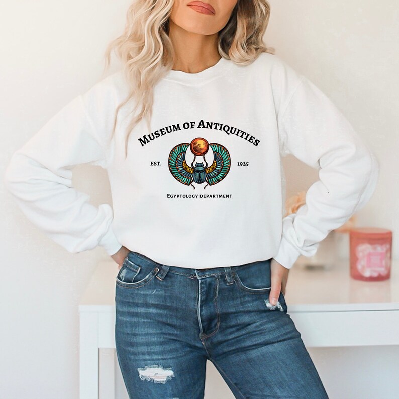 Puede incluir: Sudadera blanca con un estampado gr&aacute;fico de un escarabajo con alas y un disco solar. El texto "Museum of Antiquities Est. 1925 Egyptology Department" est&aacute; impreso debajo del gr&aacute;fico.