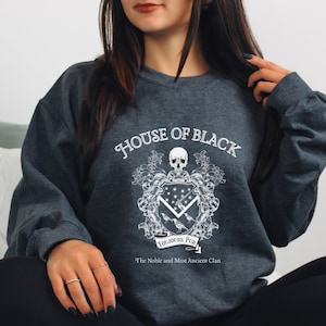 May include: A dark grey sweatshirt with a white graphic design featuring a skull, a coat of arms, and the text "House of Black, Toujours Pur, The Noble and Most Ancient Clan".