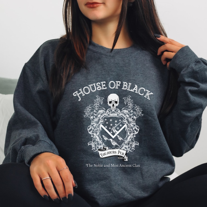 May include: A dark grey sweatshirt with a white graphic design featuring a skull, a coat of arms, and the text "House of Black, Toujours Pur, The Noble and Most Ancient Clan".
