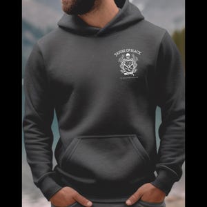 May include: Dark gray hoodie with a front pocket and a hood. The hoodie features a white graphic on the upper left chest that includes a skull, crossed swords, and the text "HOUSE OF BLACK". The background is a blurred view of a body of water and trees.