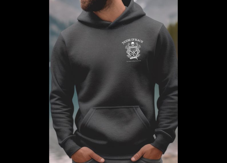 May include: Dark gray hoodie with a front pocket and a hood. The hoodie features a white graphic on the upper left chest that includes a skull, crossed swords, and the text "HOUSE OF BLACK". The background is a blurred view of a body of water and trees.
