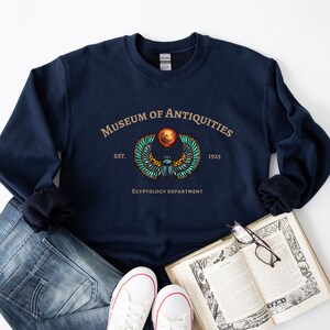 Puede incluir: Una sudadera azul marino con un dise&ntilde;o gr&aacute;fico de un escarabajo con alas y un disco solar. El texto "Museum of Antiquities Est. 1925 Egyptology Department" est&aacute; impreso en la sudadera.