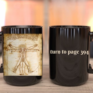 May include: Black ceramic coffee mug with a white image of the Vitruvian Man on one side and the text "turn to page 394" on the other side.