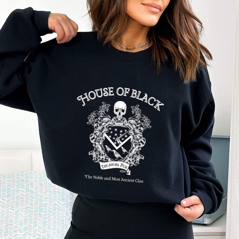 May include: Black sweatshirt featuring a white graphic of a coat of arms. The graphic includes a skull, a shield with stars and a sword, and three birds. The text "House of Black" and "Toujours Pur" are included in the graphic. The text "The Noble and Most Ancient Clan" is below the graphic.
