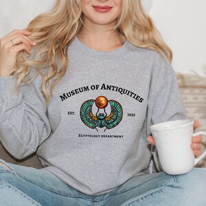Puede incluir: Sudadera gris con un gr&aacute;fico de un escarabajo con alas y un disco solar. El texto "Museum of Antiquities Est. 1925 Egyptology Department" est&aacute; impreso debajo del gr&aacute;fico.