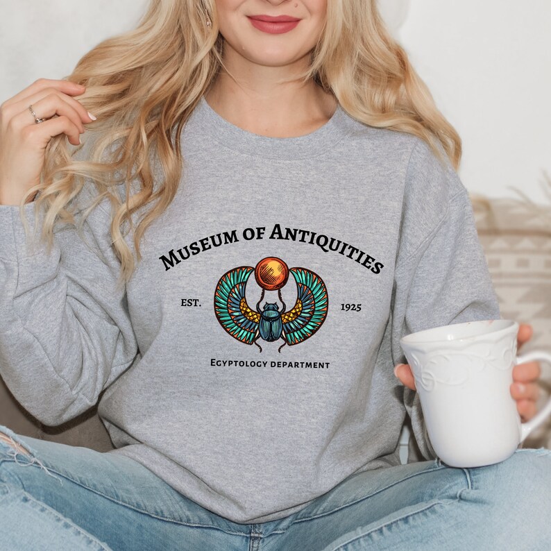 Puede incluir: Sudadera gris con un gr&aacute;fico de un escarabajo con alas y un disco solar. El texto "Museum of Antiquities Est. 1925 Egyptology Department" est&aacute; impreso debajo del gr&aacute;fico.