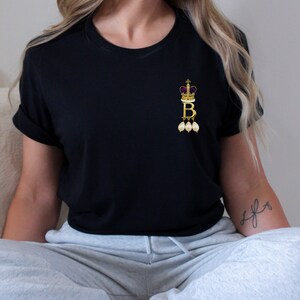 Może przedstawiać: Czarny t-shirt z złotą koroną i literą "B" z perłowymi akcentami.