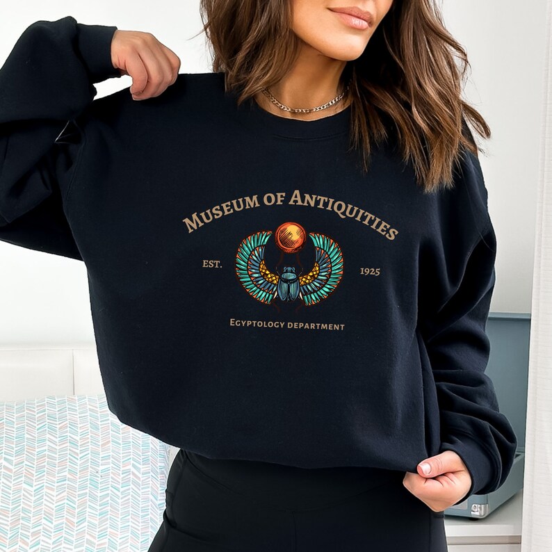 Puede incluir: Sudadera negra con un estampado gr&aacute;fico de un escarabajo egipcio con alas y un disco solar. El texto "Museum of Antiquities Est. 1925 Egyptology Department" est&aacute; impreso debajo del gr&aacute;fico.