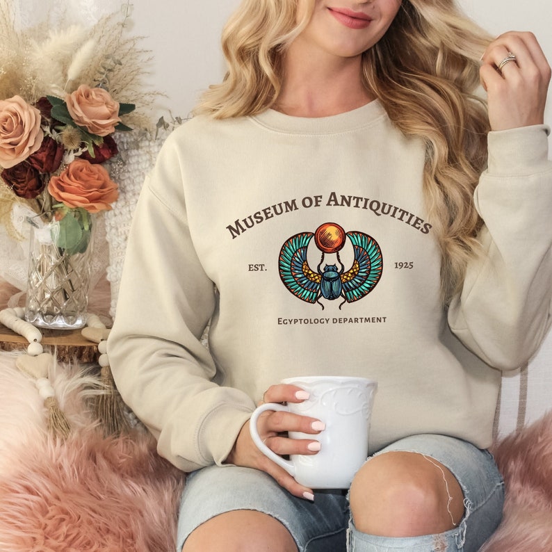 Puede incluir: Una sudadera beige con un estampado gr&aacute;fico de un escarabajo con alas y un disco solar. El texto "Museum of Antiquities Est. 1925 Egyptology Department" est&aacute; impreso debajo del gr&aacute;fico.