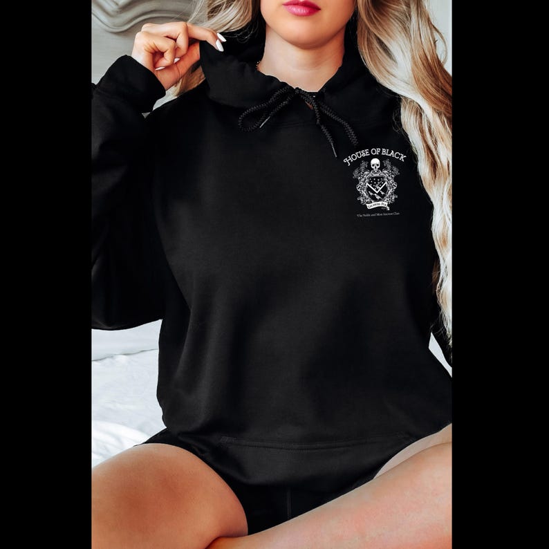 May include: Black hooded sweatshirt with a drawstring. The sweatshirt features a white graphic on the upper chest that includes a skull, floral elements, and the text "HOUSE OF BLACK". The design is centered and detailed.