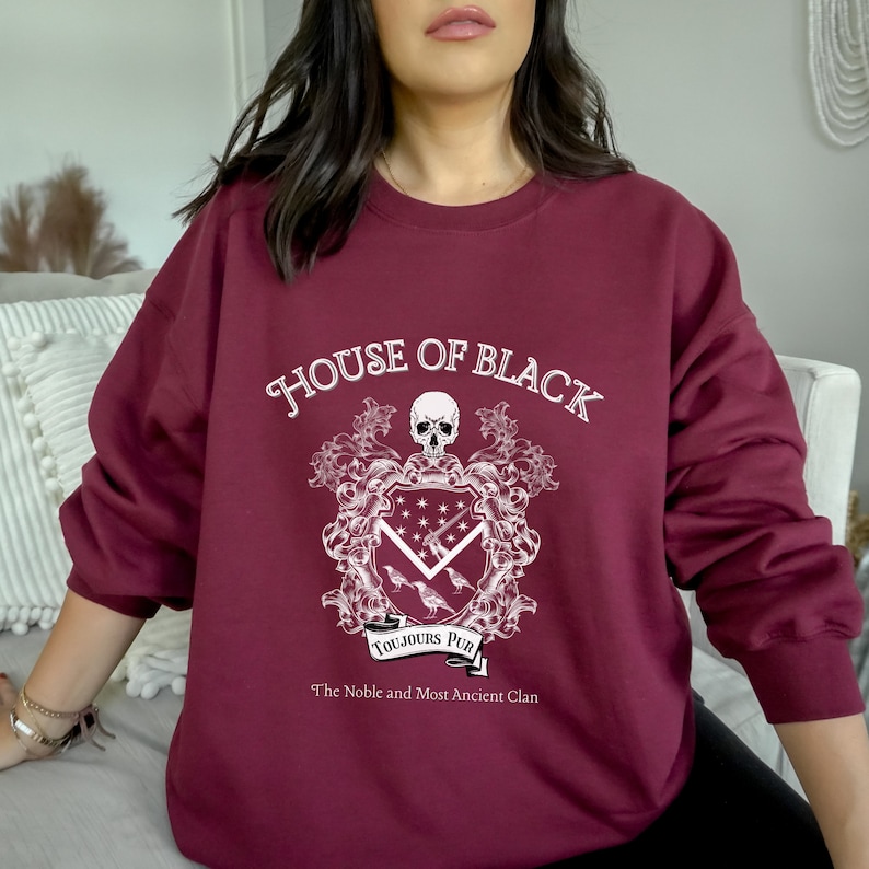 May include: A maroon sweatshirt with a white graphic design featuring a coat of arms with a skull, a banner that reads "Toujours Pur", and the text "House of Black, The Noble and Most Ancient Clan".