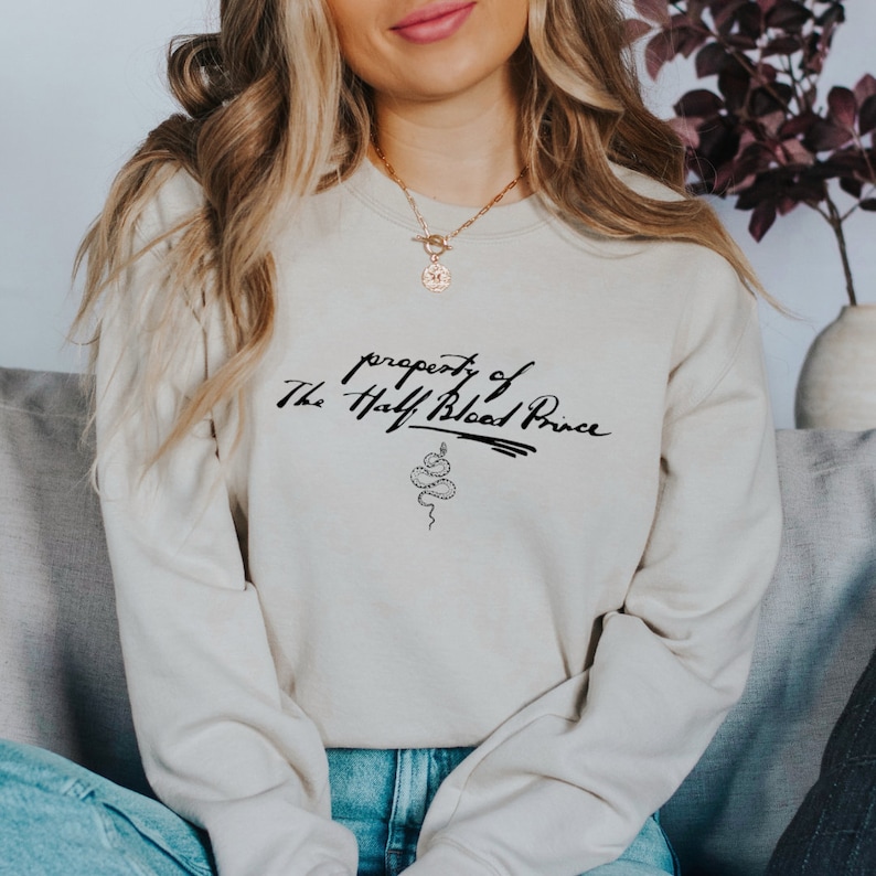 May include: A light beige sweatshirt with the text "property of The Half Blood Prince" and a snake graphic printed on the front.