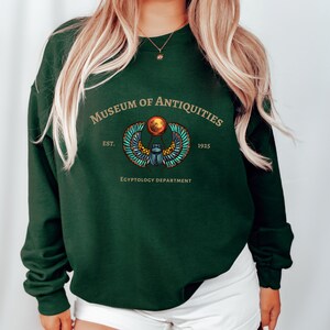 Puede incluir: Una sudadera verde oscuro con un gr&aacute;fico dorado y azul de un escarabajo egipcio alado. El texto "Museum of Antiquities Est. 1925 Egyptology Department" est&aacute; impreso en la sudadera.