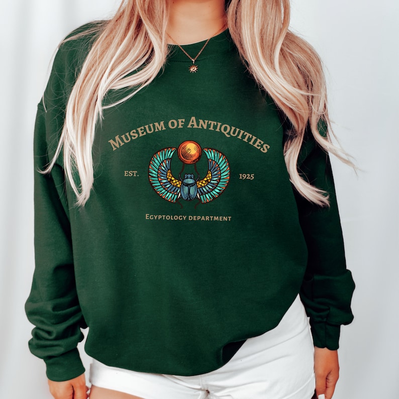 Puede incluir: Una sudadera verde oscuro con un gr&aacute;fico dorado y azul de un escarabajo egipcio alado. El texto "Museum of Antiquities Est. 1925 Egyptology Department" est&aacute; impreso en la sudadera.