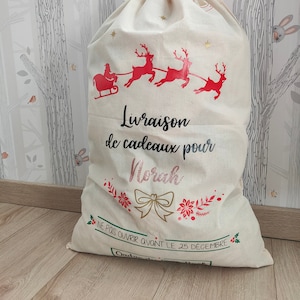 May include: A large, cream-coloured Christmas gift bag with a drawstring closure. The bag features a red illustration of Santa's sleigh pulled by reindeer, with the text "Livraison de cadeaux pour Norah" and "NE PAS OUVRIR AVANT LE 25 DECEMBRE".