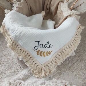 May include: A white baby blanket with a cream-colored fringe and the name "Jade" embroidered in black with a gold leaf design. The blanket is inside a woven basket with a beige blanket.
