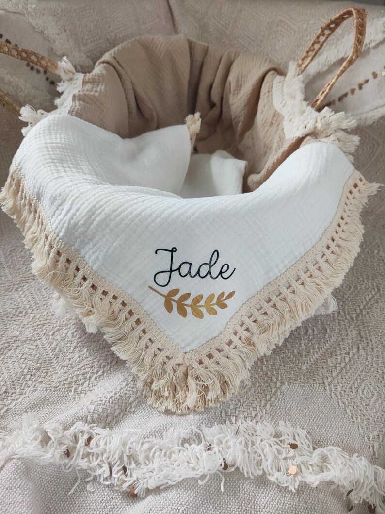 May include: A white baby blanket with a cream-colored fringe and the name "Jade" embroidered in black with a gold leaf design. The blanket is inside a woven basket with a beige blanket.