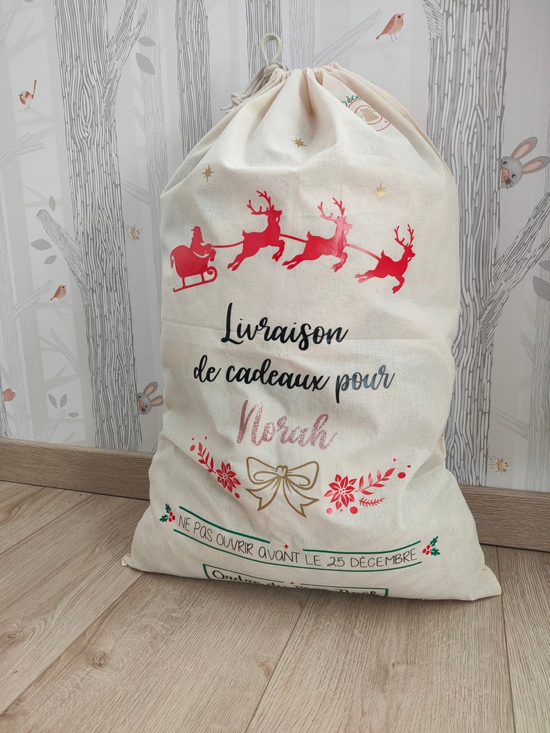 May include: A large, cream-coloured Christmas gift bag with a drawstring closure. The bag features a red illustration of Santa's sleigh pulled by reindeer, with the text "Livraison de cadeaux pour Norah" and "NE PAS OUVRIR AVANT LE 25 DECEMBRE".