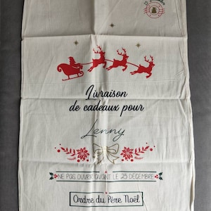 May include: A cream-coloured Christmas gift bag featuring a red Santa and reindeer design. The bag has French text, including "Livraison de cadeaux pour Lenny" and "Ne pas ouvrir avant le 25 d&eacute;cembre."