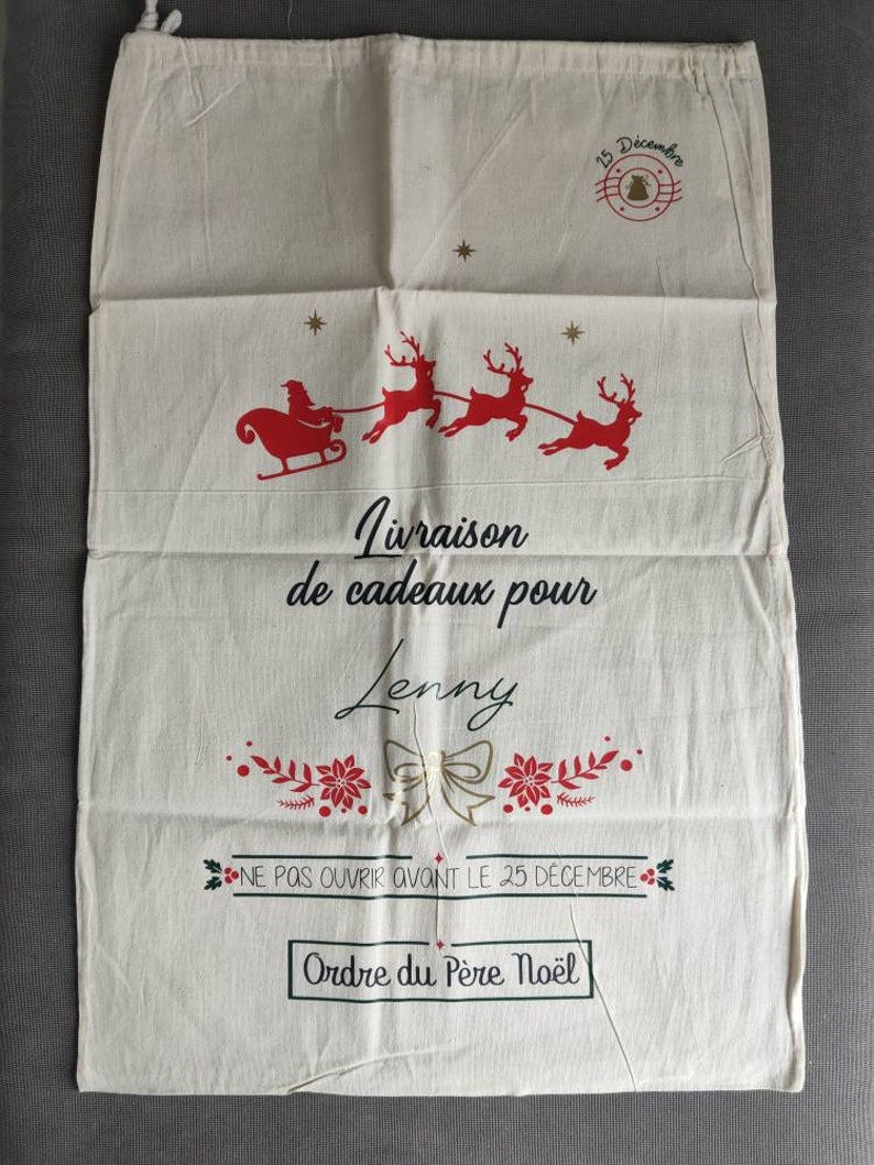 May include: A cream-coloured Christmas gift bag featuring a red Santa and reindeer design. The bag has French text, including "Livraison de cadeaux pour Lenny" and "Ne pas ouvrir avant le 25 d&eacute;cembre."