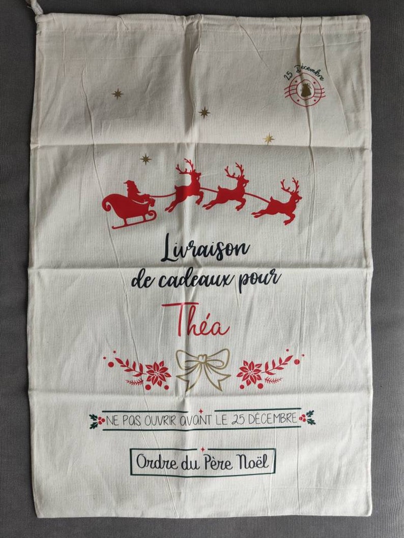 May include: Cream Christmas gift bag with red and gold details. Depicts Santa and reindeer, with text saying "Delivery of gifts for Thea" and "Do not open before December 25th". Includes a drawstring closure for securing gifts.