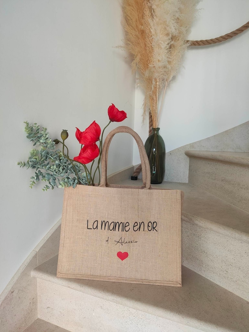 May include: A burlap tote bag with the text "La mamie en or d'Alessio" and a red heart. The bag is sitting on a set of stairs with a bouquet of red poppies and green foliage in the background.