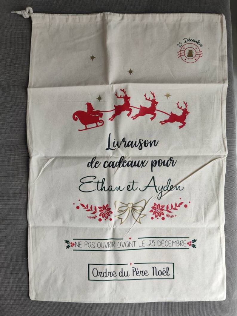 May include: A cream-coloured Christmas gift bag featuring a red Santa and reindeer design. The bag has French text: "Delivery of gifts for Ethan and Ayden" and "Do not open before December 25th."