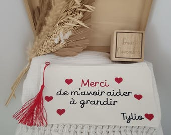 May include: A wooden box containing a white fabric pouch with a red tassel and the text "Merci de m'avoir aider à grandir" with red hearts and the name "Tylio". Inside are dried beige grasses and a small wooden block labelled "Doux Novembre".
