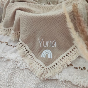 May include: Beige muslin blanket with a fringed edge. The blanket features the name "Yuna" embroidered in white, along with a white rainbow graphic. The blanket is folded and draped over a textured surface.