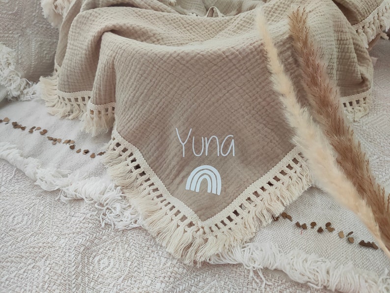 May include: Beige muslin blanket with a fringed edge. The blanket features the name "Yuna" embroidered in white, along with a white rainbow graphic. The blanket is folded and draped over a textured surface.