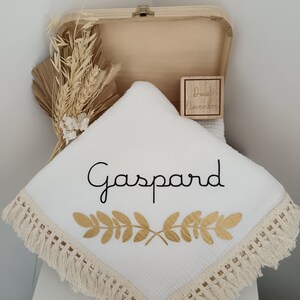 May include: A white baby blanket with the name "Gaspard" in black script and gold leaf design. The blanket has a fringe trim and is displayed in an open wooden box with dried floral accents and a wooden block.