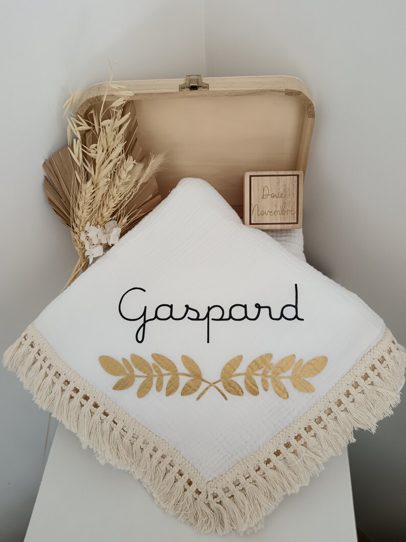 May include: A white baby blanket with the name "Gaspard" in black script and gold leaf design. The blanket has a fringe trim and is displayed in an open wooden box with dried floral accents and a wooden block.