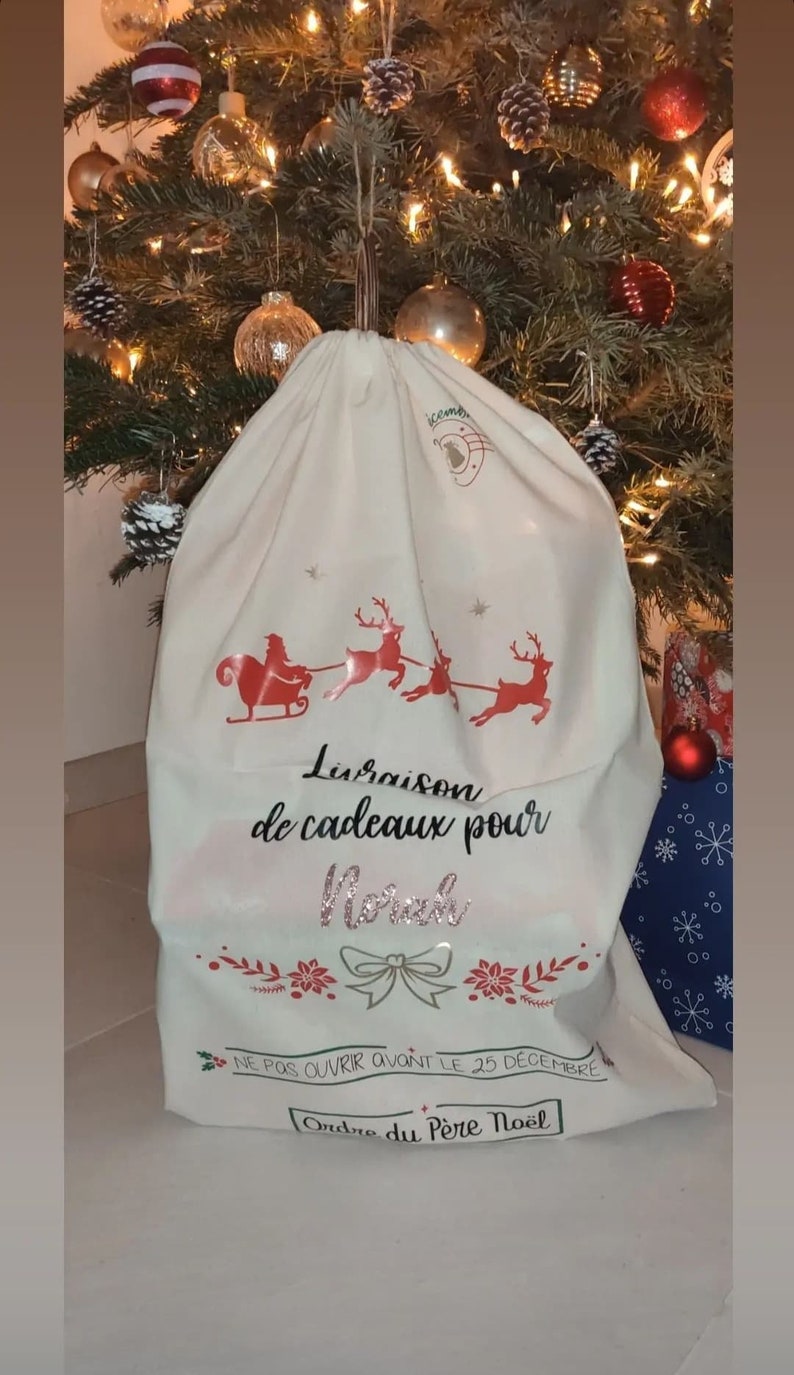 May include: A cream Christmas gift bag featuring a red Santa and reindeer design. The bag has text that reads "Delivery of gifts for Norah" and "Do not open before December 25th".