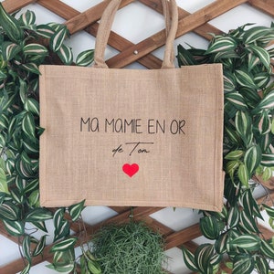 May include: A brown burlap tote bag with the text "Ma Mamie en Or de Ton" and a red heart. The bag is hanging on a wooden lattice.