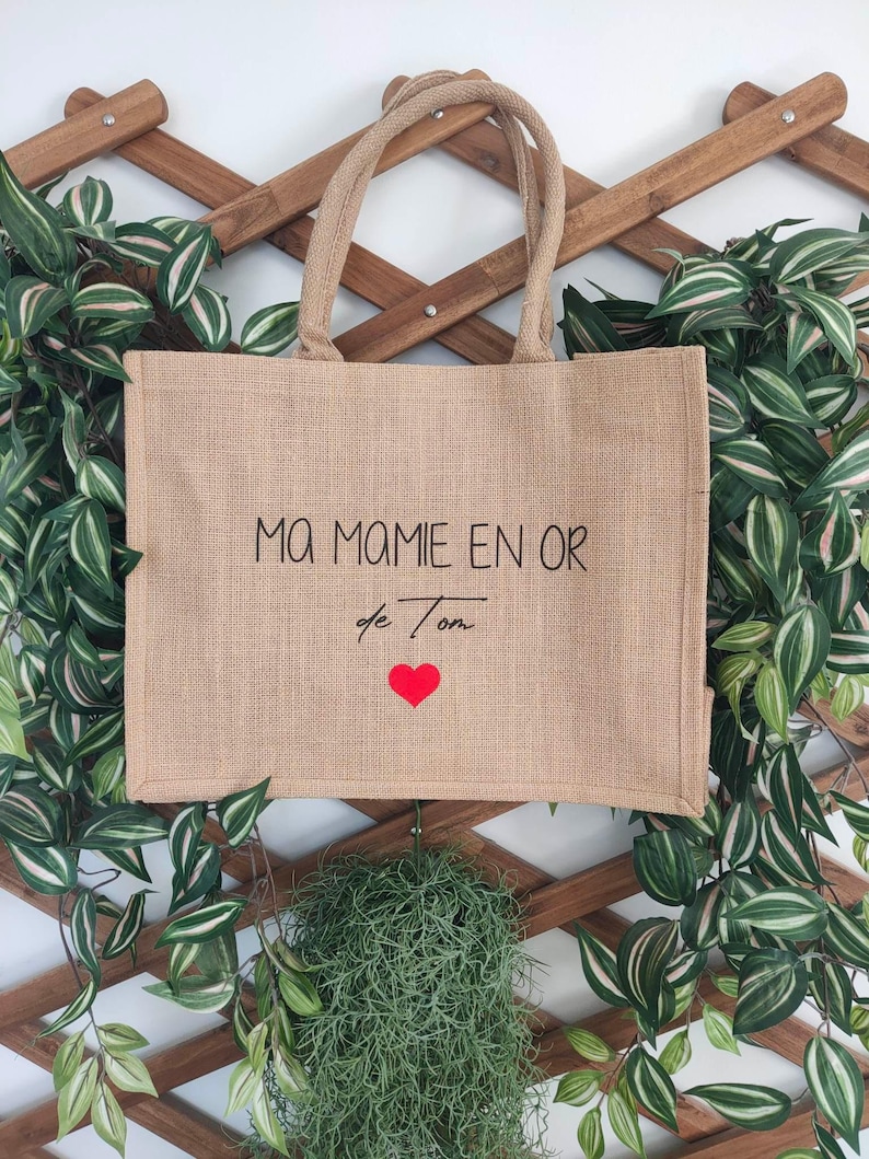 May include: A brown burlap tote bag with the text "Ma Mamie en Or de Ton" and a red heart. The bag is hanging on a wooden lattice.