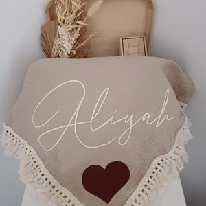 May include: A beige baby blanket with the name "Aliyah" in white script, a brown heart, and a fringed edge. The blanket is displayed with a wooden box, dried flowers, and a small wooden block with text.