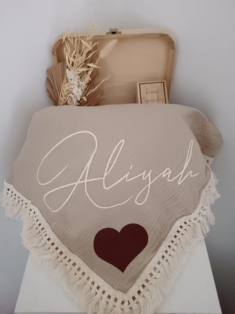 May include: A beige baby blanket with the name "Aliyah" in white script, a brown heart, and a fringed edge. The blanket is displayed with a wooden box, dried flowers, and a small wooden block with text.