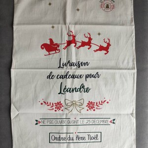 May include: A cream-coloured Christmas gift bag with a drawstring closure. The bag features a red illustration of Santa's sleigh and reindeer, along with the text "Delivery of gifts for Leandro" and other festive text.