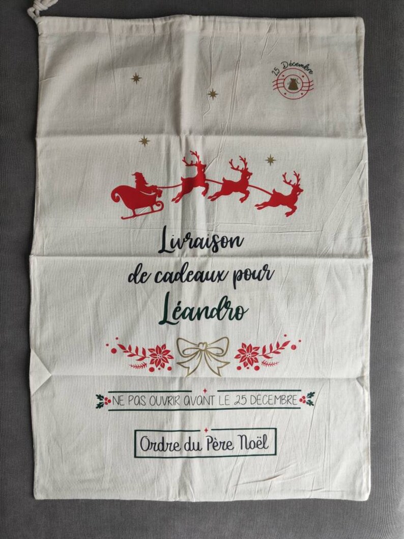 May include: A cream-coloured Christmas gift bag with a drawstring closure. The bag features a red illustration of Santa's sleigh and reindeer, along with the text "Delivery of gifts for Leandro" and other festive text.