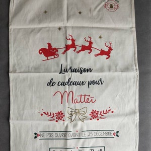 May include: A cream-coloured Christmas gift bag with a drawstring closure. The bag features a red illustration of Santa's sleigh and reindeer, along with text in French. The text includes "Livraison de cadeaux pour Matte&iuml;" and "Ne pas ouvrir avant le 25 d&eacute;cembre."