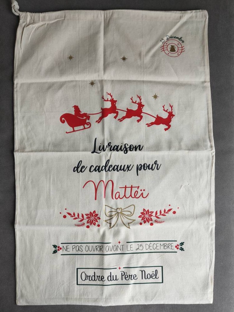 May include: A cream-coloured Christmas gift bag with a drawstring closure. The bag features a red illustration of Santa's sleigh and reindeer, along with text in French. The text includes "Livraison de cadeaux pour Matte&iuml;" and "Ne pas ouvrir avant le 25 d&eacute;cembre."