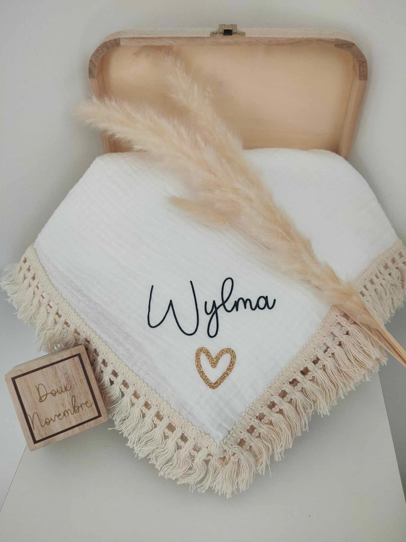 May include: A white baby blanket with a cream-colored fringe and the name "Wylma" in black script, along with a gold glitter heart. A wooden box and pampas grass are also in the image.