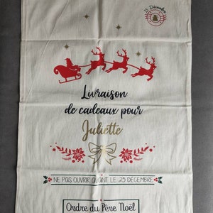 May include: A cream Christmas gift bag with a drawstring. It features a red Santa and reindeer design, with the text "Livraison de cadeaux pour Juliette" and "Ne pas ouvrir avant le 25 d&eacute;cembre". The bag is decorated with festive imagery.
