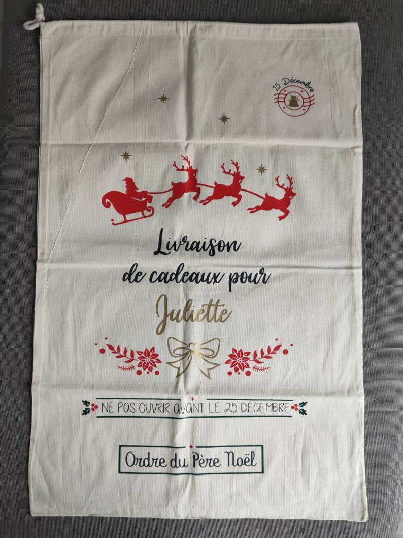 May include: A cream Christmas gift bag with a drawstring. It features a red Santa and reindeer design, with the text "Livraison de cadeaux pour Juliette" and "Ne pas ouvrir avant le 25 d&eacute;cembre". The bag is decorated with festive imagery.