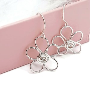 May include: Silver flower dangle earrings with a twisted centre. The earrings are hanging from a pink box.